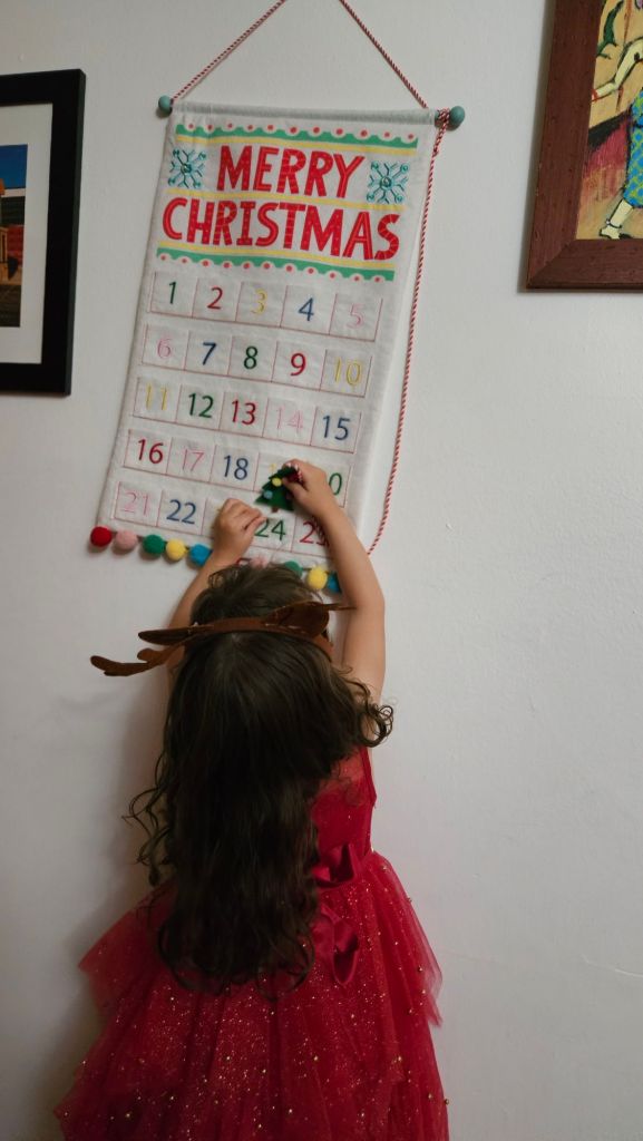A little girl wearing a red Christmas dress puts a green, felt Christmas tree in a pocket labeled "24" on an Advent calendar.